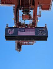 A shipping container with a United States flag banner hangs from a crane at Terminal 46 in Seattle.