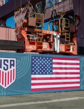 A shipping container with a United States flag banner at Terminal 46 before it is hung from a crane. 