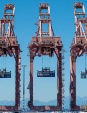 Three containers hang from cranes at Terminal 46 in Seattle. The middle container has a “Welcome to the Pacific Northwest” message to FIFA World Cup visitors, while the outside containers have a Bosnia-Herzegovina and Qatar flag.