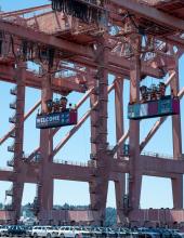 Three containers hang from cranes at Terminal 46 in Seattle. The middle container has a “Welcome to the Pacific Northwest” message to FIFA World Cup visitors, while the outside containers have Bosnia-Herzegovina and Qatar flags.
