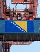 A shipping container with a Bosnia and Herzegovina flag banner hangs from a crane at Terminal 46 in Seattle.