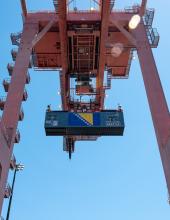 A shipping container with a Bosnia and Herzegovina flag banner hangs from a crane at Terminal 46 in Seattle.