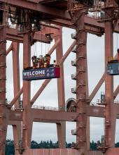 Three containers hang from cranes at Terminal 46 in Seattle. The middle container has a “Welcome to the Pacific Northwest” message to FIFA World Cup visitors, while the outside containers have the Egypt and IR Iran flags.