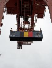 A shipping container with a Belgium flag banner hangs from a crane at Terminal 46 in Seattle.