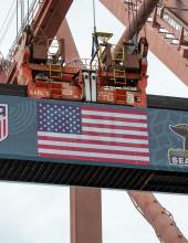A shipping container with a United States flag banner hangs from a crane at Terminal 46 in Seattle.